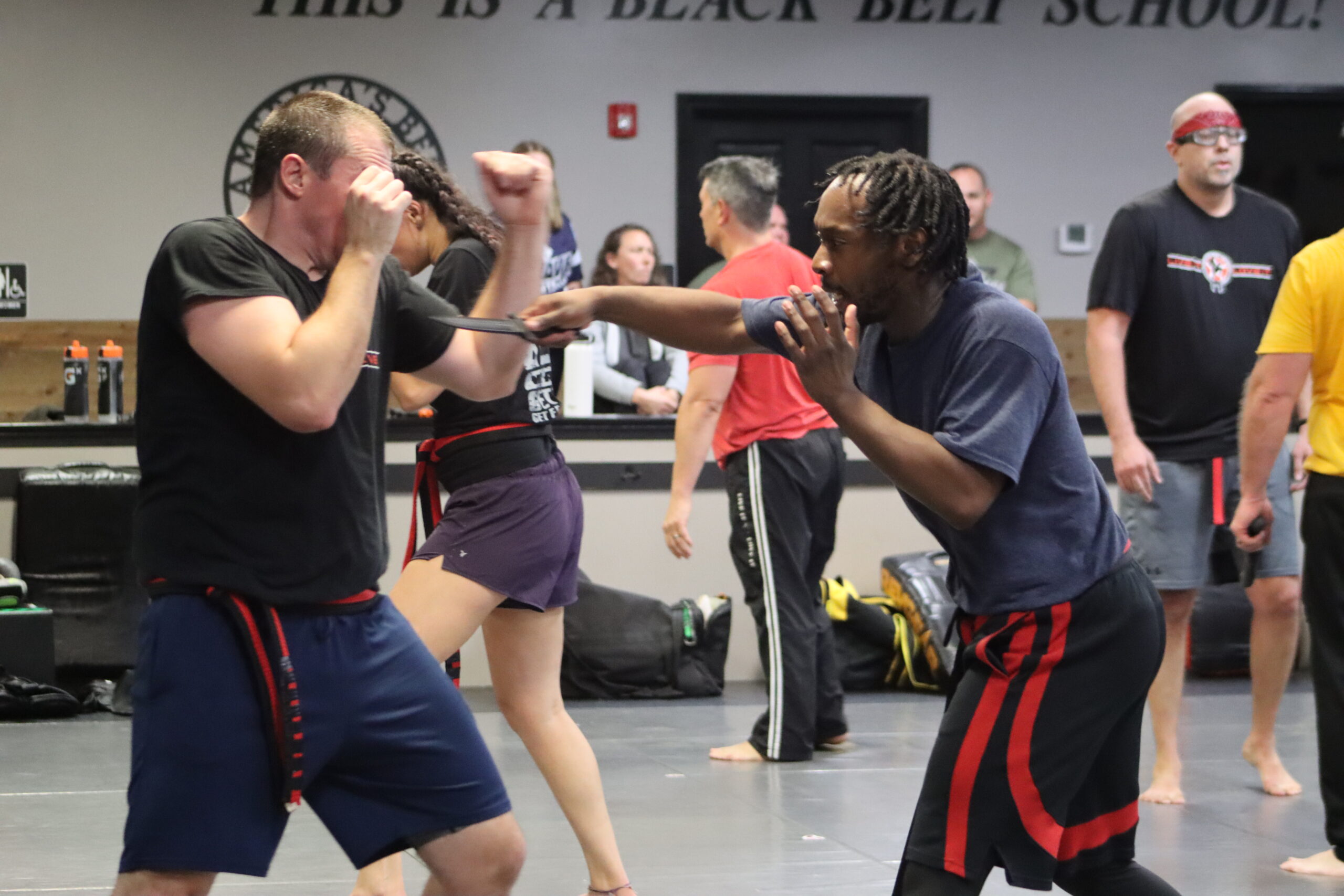 Students practicing a self-defense drill during a Krav Maga class at America's Best Defense. One student is simulating an attack with a training weapon while the other defends, demonstrating focus and technique. Other participants and instructors observe and train in the background in a dynamic and engaging class environment.
