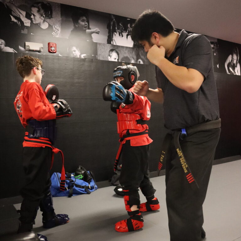Martial arts instructor wearing black polo, black gi pants and black belt works with two young students wearing red gi tops, black pants, and red and black belts.