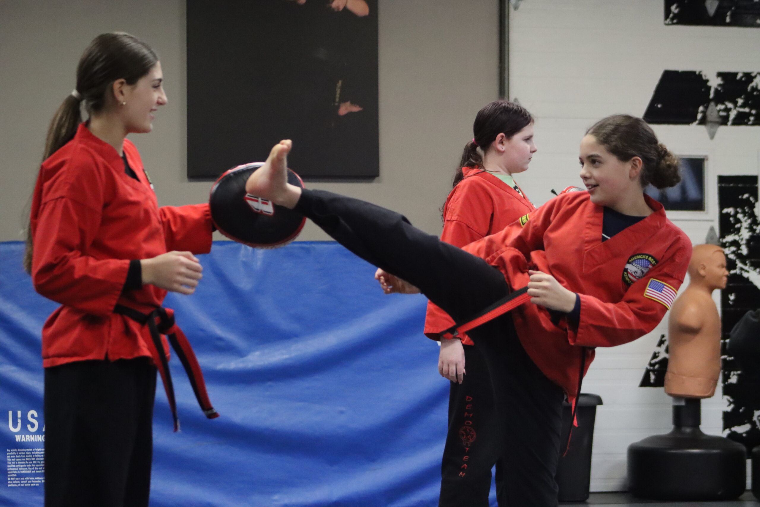 Preteen girls wearing red gi tops, black pants, and red and black belts practice martial arts in a dojo.