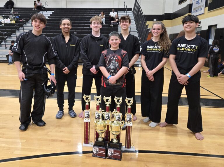 Team Next Element martial arts competitors, including ABD Franklin students Gregory Perella and Connor Poirier, posing together in a gym with multiple trophies in front of them.