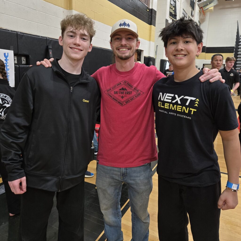 Gregory Perella, Sensei Mike Saunders, and Connor Poirier smiling and posing together in a gymnasium after a martial arts competition.
