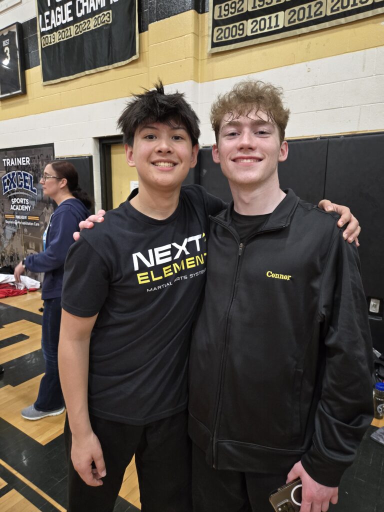 ABD Franklin students Gregory Perella and Connor Poirier smiling and posing together in a gymnasium, wearing Team Next Element gear after a martial arts competition