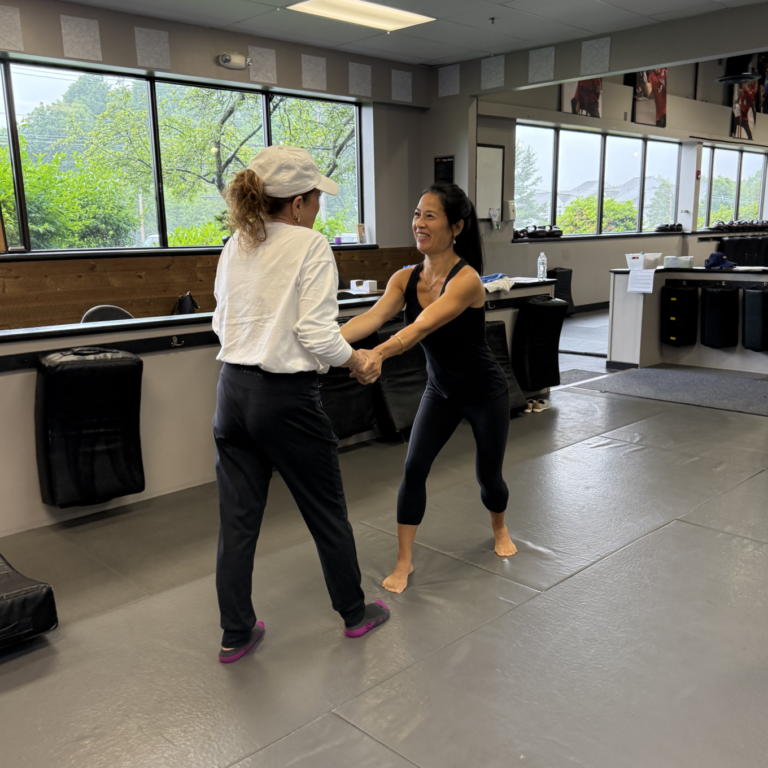 A woman with dark hair wearing a black tank top and black leggings is grabbing the arms of a woman with a ponytail wearing black leggings, a white top, and a light-colored hat. The women are in a martial arts setting.