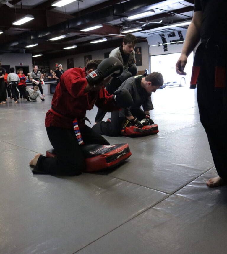 Young martial artists wearing red, black, and gray uniforms hit pad on the ground at America's Best Defense.